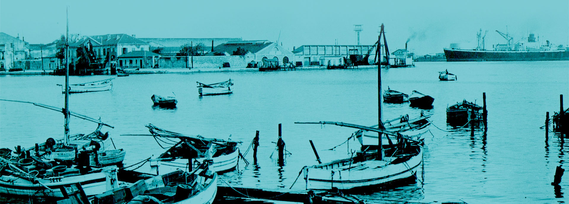 Des bateaux de pêche traditionnels sont amarrés dans le port, avec des filets et des paniers visibles à bord. En arrière-plan, des bâtiments industriels et des entrepôts longent le quai, donnant une idée de l'activité maritime et commerciale de l'époque. Un grand navire de fret est visible au loin, soulignant l'importance du port pour le commerce international. L'image est teintée en bleu, ce qui ajoute une touche nostalgique et met en valeur le contraste entre les éléments maritimes et industriels.